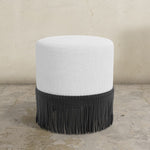 Round upholstered ottoman with white top and black fringed bottom on a textured floor.