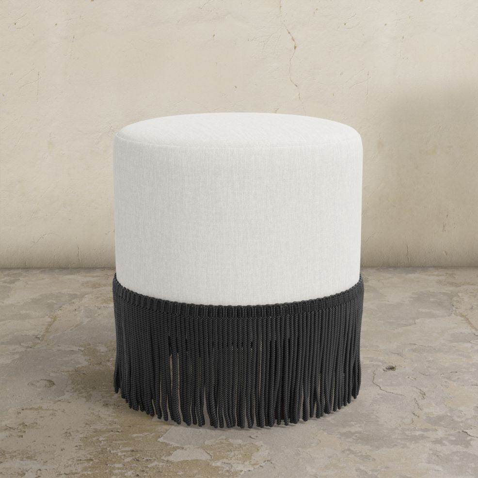 Round upholstered ottoman with white cushion and fringes on a concrete floor.
