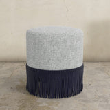 Round upholstered ottoman with gray top and navy fringes on a concrete floor.