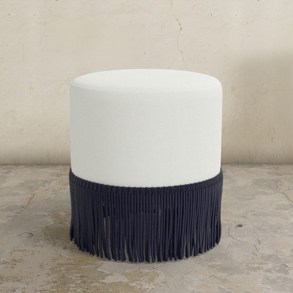 Round upholstered ottoman with white top and navy fringes on a concrete floor.