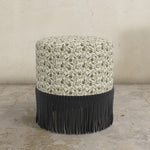Round upholstered ottoman with floral pattern and black fringe on a concrete floor.