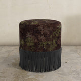 Round upholstered ottoman with rich floral pattern and fringed edge on a concrete floor.
