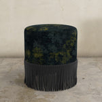 Floral-patterned upholstered ottoman with fringe on a textured floor and light-colored wall background