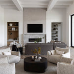 A modern living room with a large round black coffee table surrounded by four textured light beige armchairs on a woven gray and cream area rug. In the background, a gray sectional sofa sits in front of a fireplace with a mounted TV above it, under a white coffered ceiling.