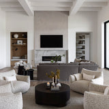 A modern living room with a large round black coffee table surrounded by four textured light beige armchairs on a woven gray and cream area rug. In the background, a gray sectional sofa sits in front of a fireplace with a mounted TV above it, under a white coffered ceiling.