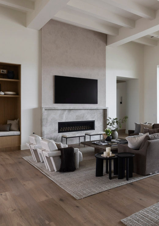 A modern living room interior featuring a large black flat-screen TV mounted above a stone fireplace. The room has light wood floors, a textured gray and cream area rug, a gray sofa with pillows, white accent chairs, and a built-in wooden shelf on the left.