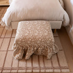 The Eloise Ruffle Ottoman in patterned fabric with solid wood construction at the base of a bed with a run underneath