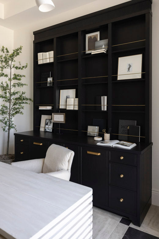 White wooden unique desk and black bookshelf with decorative items in a home workspace with a chair and plant.